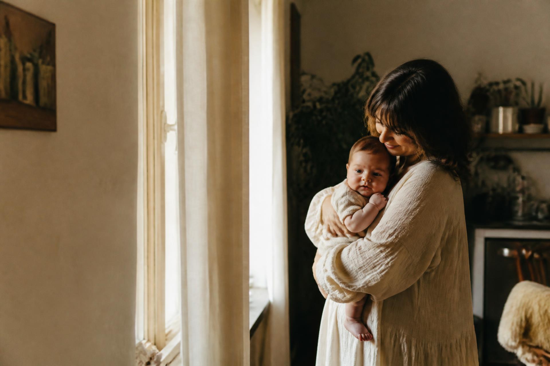 A mother holding her newborn beside a sunlit window at home